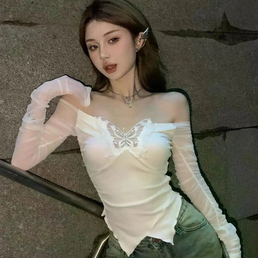 Woman wearing a white lace top and green pants against a gray wall.