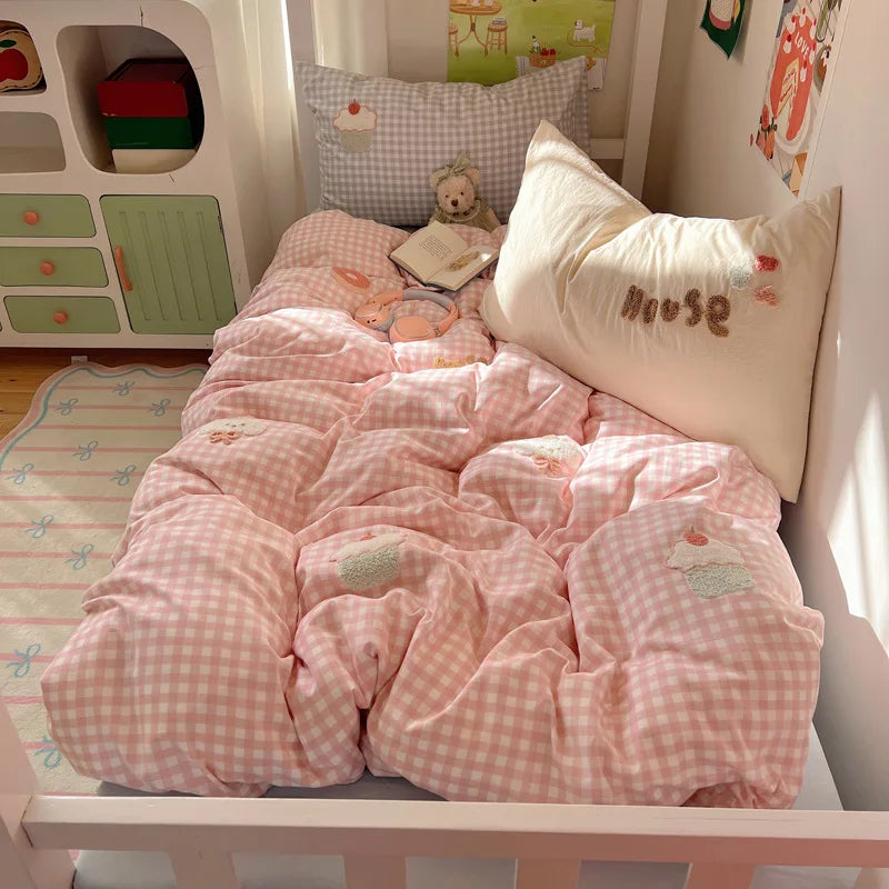 A pink and white gingham checkered bedding set, including a comforter and matching pillowcases, styled on a child's bed in a brightly lit nursery.