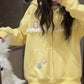 A person takes a mirror selfie wearing a pastel yellow zip-up hoodie with white lace trim, small cat and star graphics, and a front pocket. The photo is taken indoors, with a white fluffy object partially visible on the left.