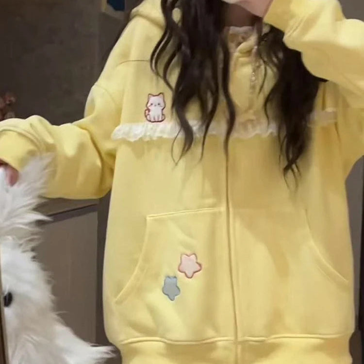 A person takes a mirror selfie wearing a pastel yellow zip-up hoodie with white lace trim, small cat and star graphics, and a front pocket. The photo is taken indoors, with a white fluffy object partially visible on the left.
