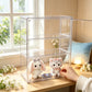 A clear acrylic shoe organizer box with three shelves stands on a wooden table in a bright room. Two small plush toys sit inside the organizer, and a person's hand is reaching in to adjust them.