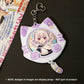 A white cat-shaped keychain holder with purple paw-print accents and a circular anime character badge insert, shown on a dark surface with a printed card partially visible at the left edge. A note at the bottom states that the badges shown are display props and not part of the product.