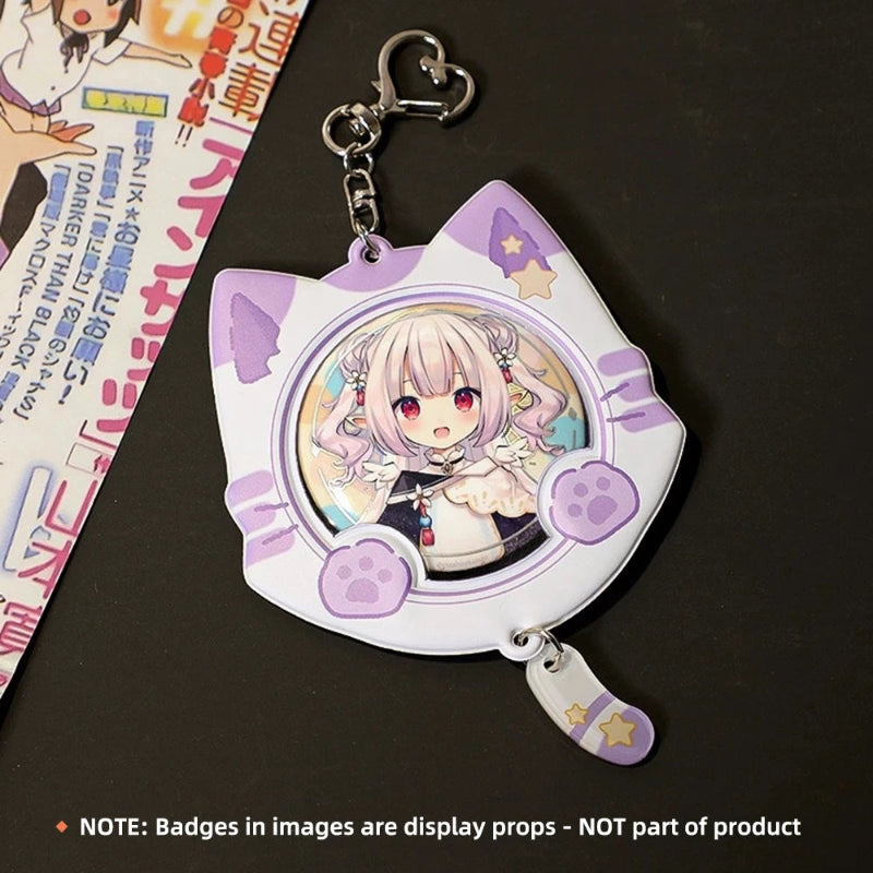 A white cat-shaped keychain holder with purple paw-print accents and a circular anime character badge insert, shown on a dark surface with a printed card partially visible at the left edge. A note at the bottom states that the badges shown are display props and not part of the product.