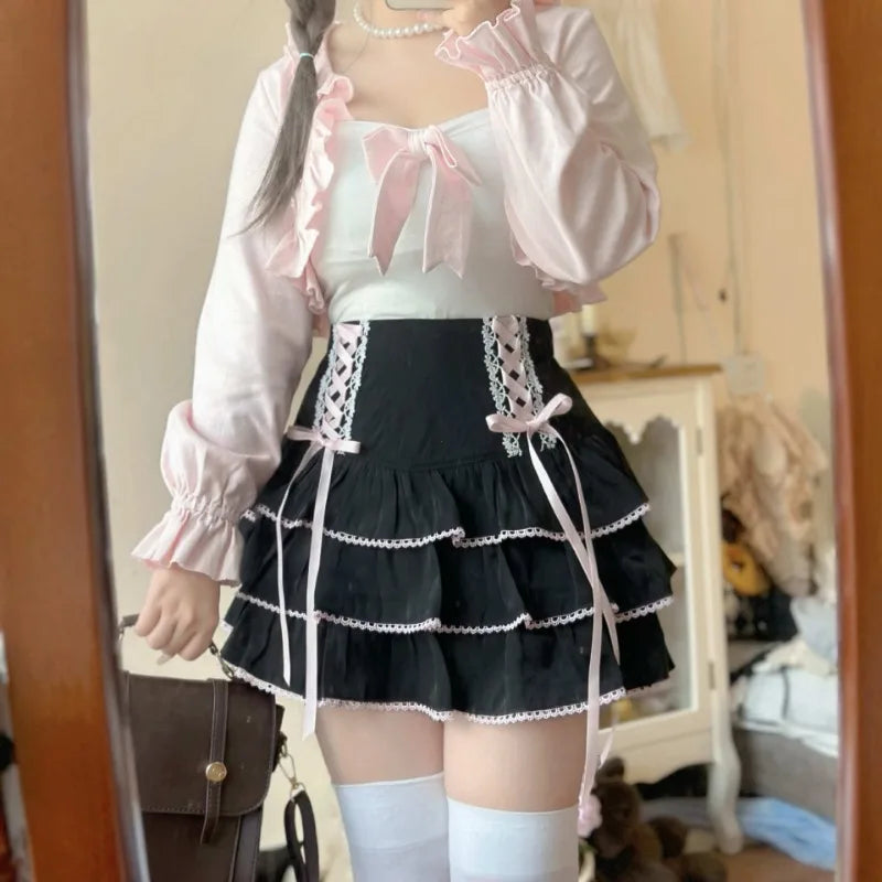 A person wearing a pink ruffled long-sleeve crop top over a white camisole and a black tiered ruffle skirt with white lace trim and ties, standing indoors in front of a mirror.