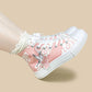 A pair of pink high-top canvas sneakers with white laces, white rubber soles, and white cloud patterns, featuring a silver crane graphic on the side. One shoe is shown being worn with a white ruffled sock against a solid beige background.