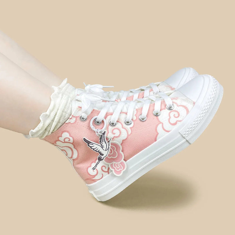 A pair of pink high-top canvas sneakers with white laces, white rubber soles, and white cloud patterns, featuring a silver crane graphic on the side. One shoe is shown being worn with a white ruffled sock against a solid beige background.