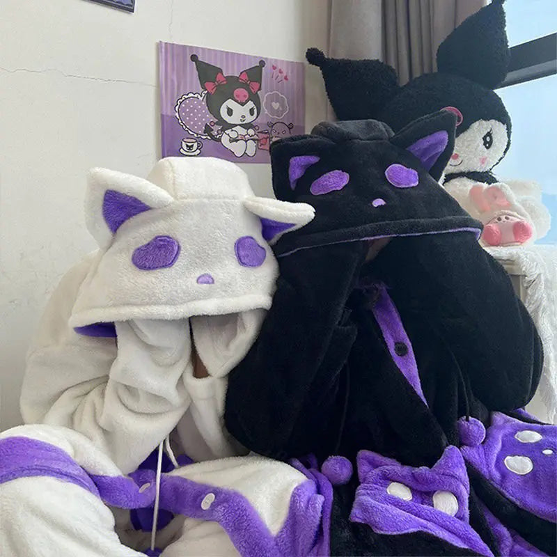 Two people wearing plush hooded cat-themed sleepwear or robes, one white with purple accents and one black with purple accents, sitting indoors with Kuromi wall art and a plush figure in the background.