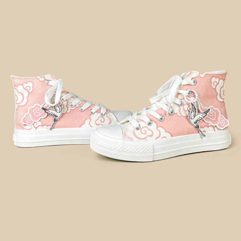 A pair of high-top canvas sneakers in light pink with white cloud patterns and silver crane charms, shown on a solid beige background.