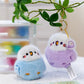 Two cute bird keychains, one blue and one purple, hanging from a branch with a blurred indoor background.