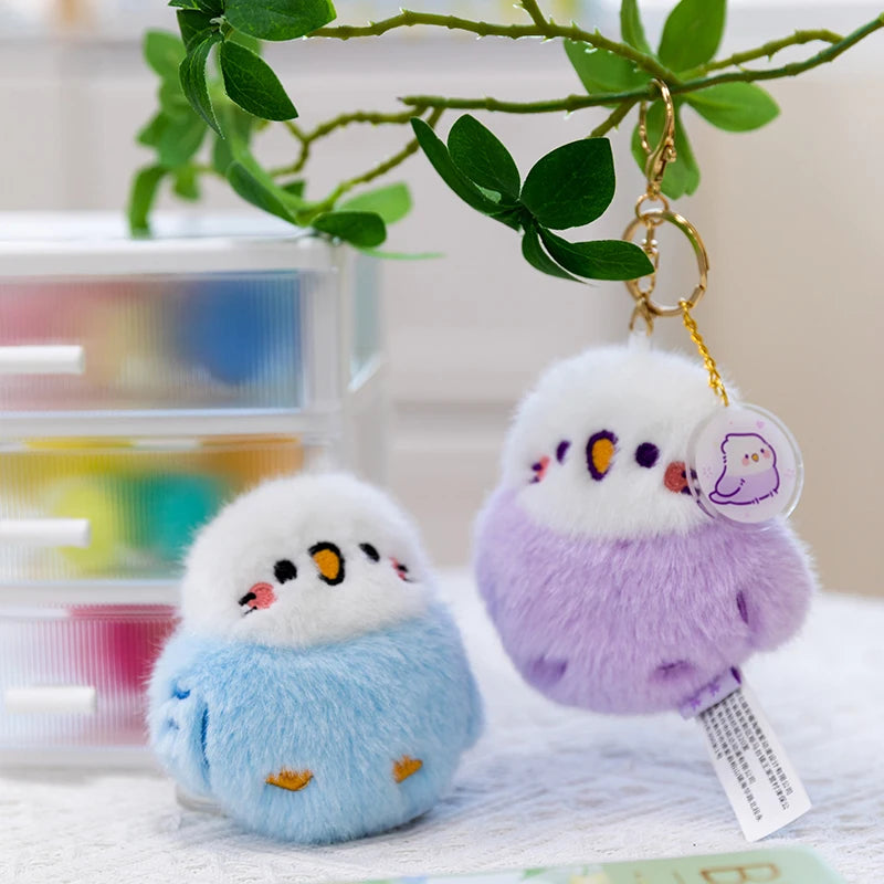 Two cute bird keychains, one blue and one purple, hanging from a branch with a blurred indoor background.