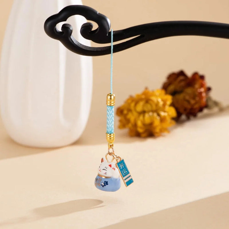 A decorative hanging charm with a small blue-and-white lucky cat pendant, a light blue braided cord, gold-tone beads, and a small hanging tag, displayed on a black curved stand against a warm beige background with a white vase and dried flowers.