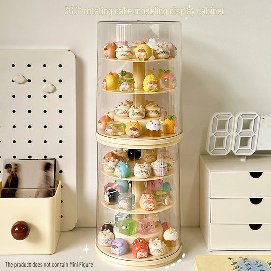 A 360-degree rotating cake modeling display cabinet with a clear cylindrical top and beige wooden tiers, filled with small decorative cupcakes and character figurines, shown on a tabletop with room decor in the background.