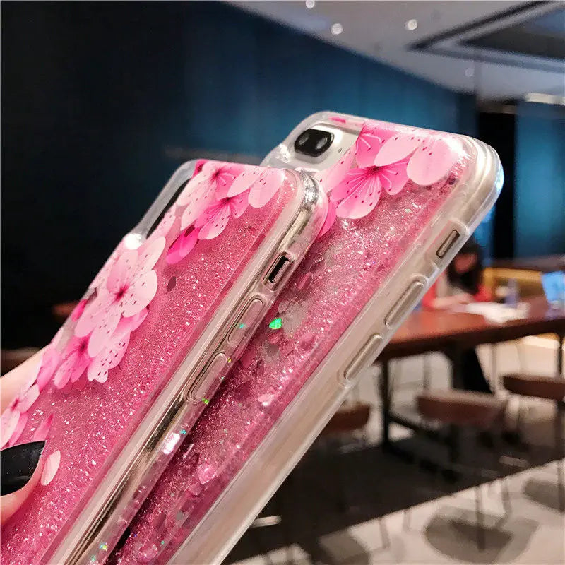 Phone case with pink cherry blossom design and glittery details, held in a hand with a blurred indoor background.