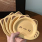 A hand holds a stack of four small, cat-shaped ceramic plates in a warm yellow color with brown ear details and simple facial features, set against a wooden table background.
