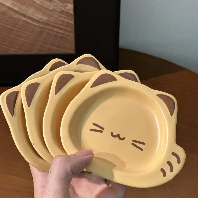 A hand holds a stack of four small, cat-shaped ceramic plates in a warm yellow color with brown ear details and simple facial features, set against a wooden table background.