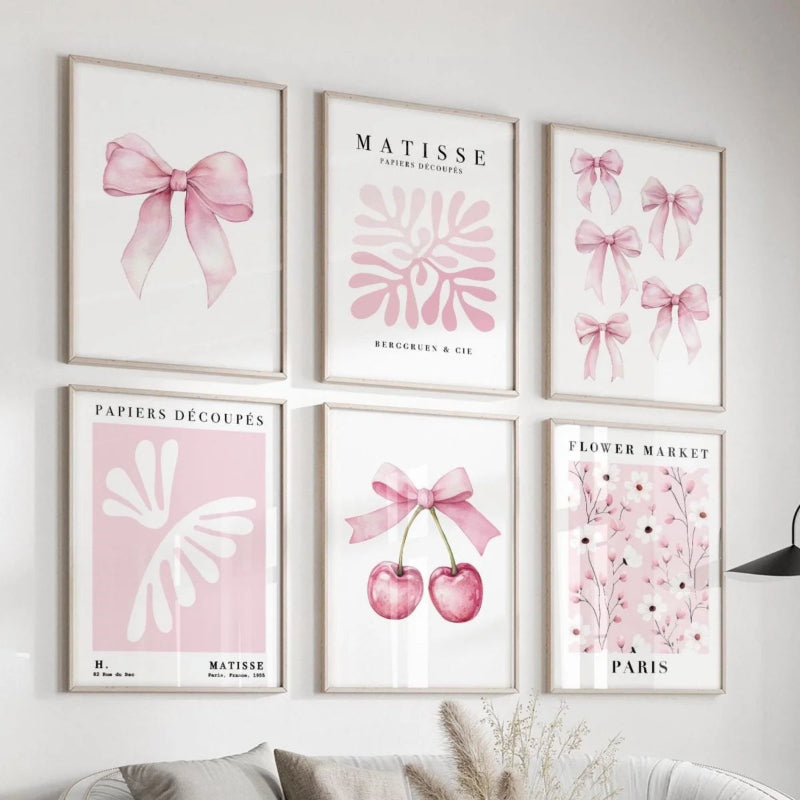 Six framed pastel art prints arranged in a gallery wall above a sofa. The prints feature pink bows, botanical motifs, cherries, and floral text including 'MATISSE' and 'PAPIERS DECOUPEES'.