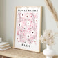 A framed floral art print leaning on a woven table against a light gray wall, with dried grasses and a white vase nearby. The print reads “FLOWER MARKET A PARIS” and features pink, white, and dark red blossoms on a pale pink background.