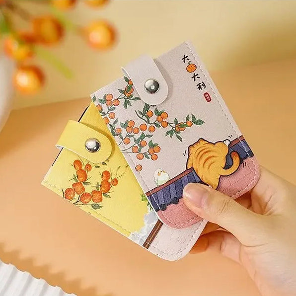 A hand holding two small decorative card cases or wallet-style phone cases with snap closures. One case is pale pink with orange fruit illustrations and a small character print, and the other is pale yellow with similar orange fruit illustrations.