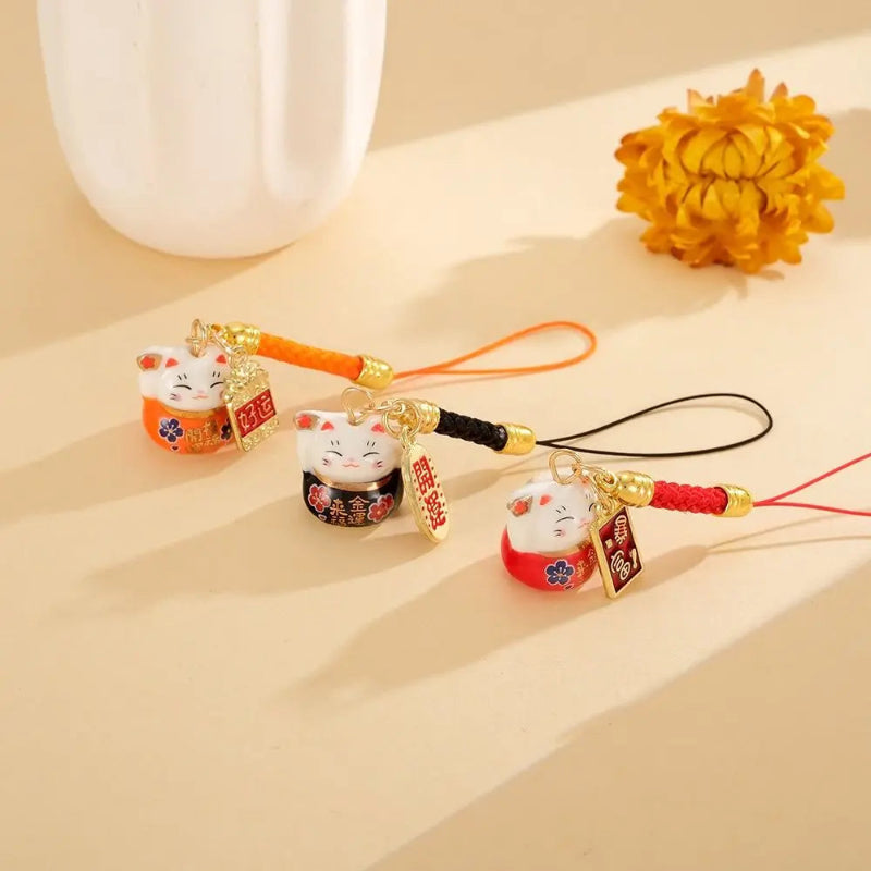 Three small lucky cat keychains or phone strap charms with white faces and colorful bodies are arranged on a warm beige surface. Each charm has a different colored cord loop (orange, black, red), gold-toned hardware, and a hanging red tag.
