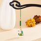 A green and gold Chinese lucky cat car charm with a hanging tag, suspended from a black decorative stand against a warm beige background with a white vase and blurred yellow flowers.
