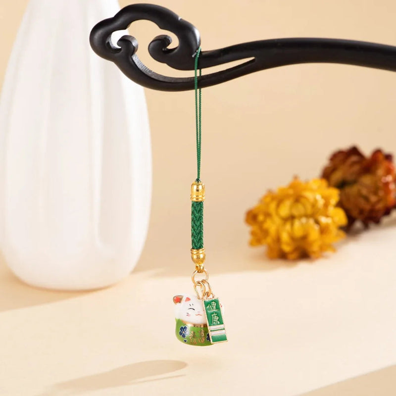 A green and gold Chinese lucky cat car charm with a hanging tag, suspended from a black decorative stand against a warm beige background with a white vase and blurred yellow flowers.