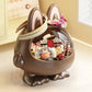 A brown bunny-shaped decorative storage container with a clear oval front window, filled with assorted small accessories and candies. The container has upright ears with pink inner linings, a bow on top, and stands on short feet on a light tabletop in a softly lit indoor setting.