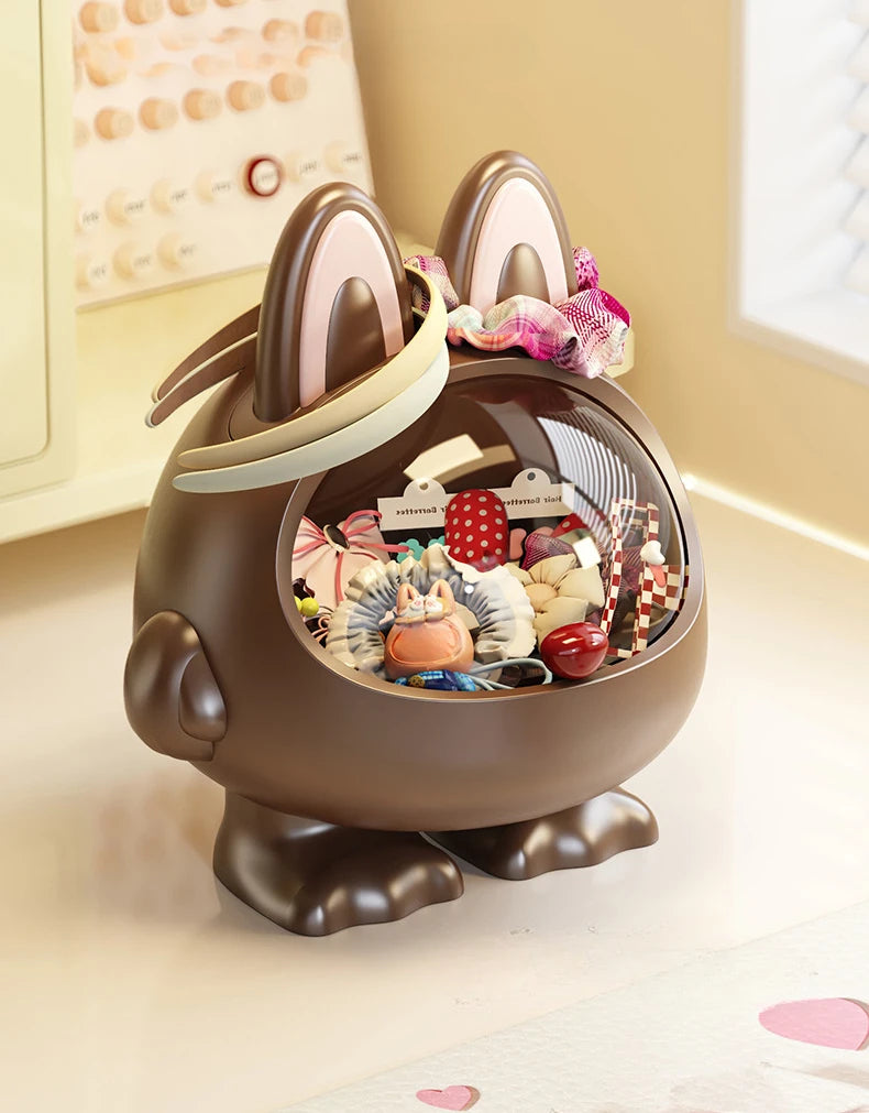 A brown bunny-shaped decorative storage container with a clear oval front window, filled with assorted small accessories and candies. The container has upright ears with pink inner linings, a bow on top, and stands on short feet on a light tabletop in a softly lit indoor setting.