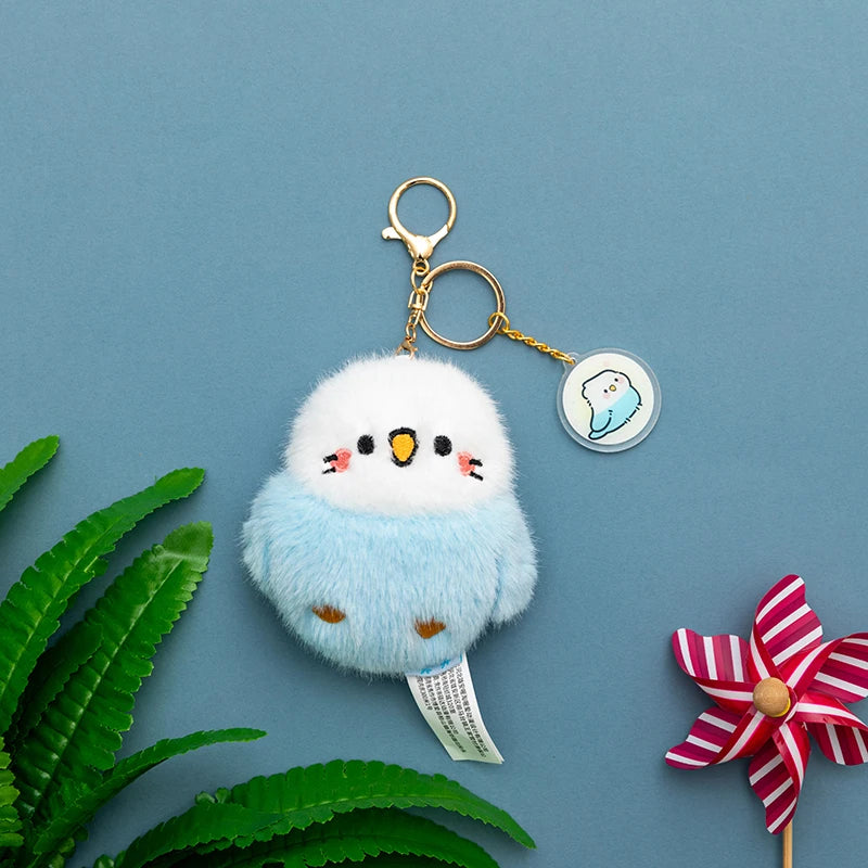 Kawaii blue and white bird plushie  keychain on a blue background with green leaves and a red and white pinwheel.