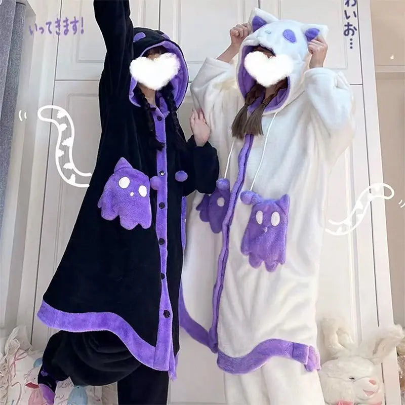 Two people wearing plush hooded kigurumi pajamas in two colorways: one black with purple trim and cat-face hood, and one white with purple trim and cat-ear hood. Both have purple ghost-shaped pocket details.