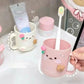 Pink kawaii mug with a toothbrush inside, next to a tube of toothpaste and a bottle on a white surface.