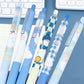 Six cute cartoon ducky-style retractable pens in pastel blue, white, and yellow tones, arranged diagonally on a blue desk with a keyboard partially visible in the background.
