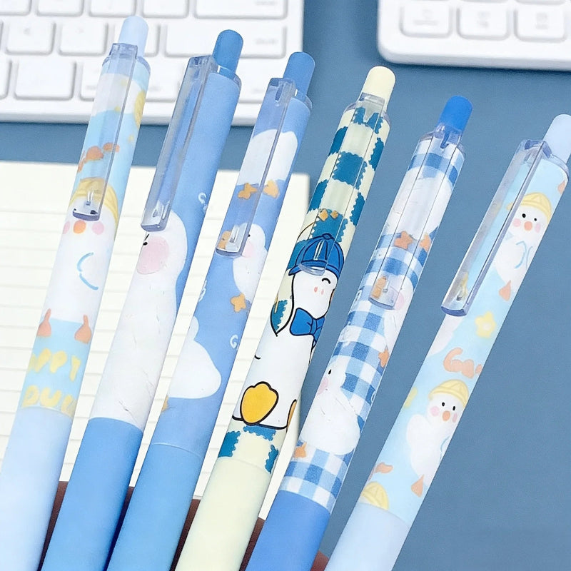 Six cute cartoon ducky-style retractable pens in pastel blue, white, and yellow tones, arranged diagonally on a blue desk with a keyboard partially visible in the background.