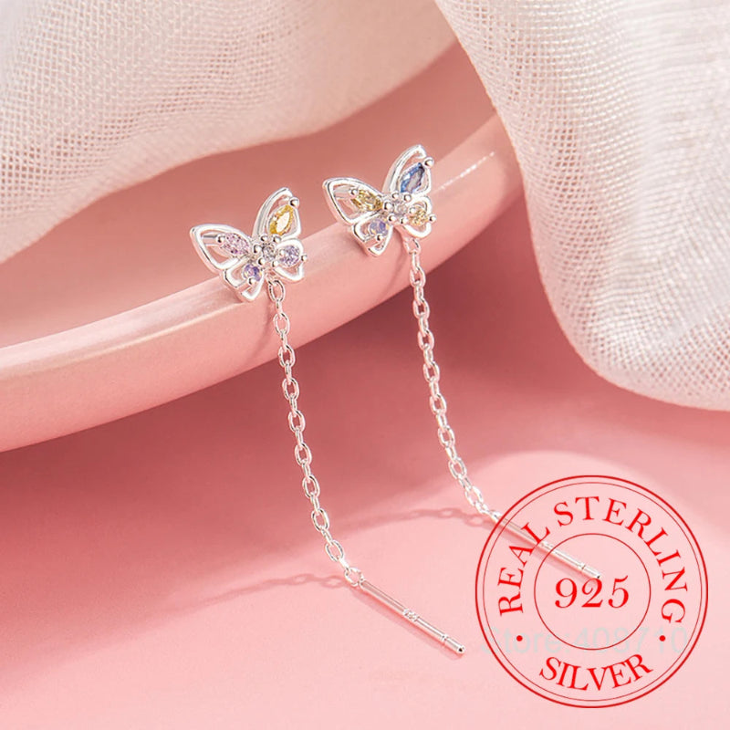 A pair of silver butterfly drop earrings with colorful gemstone accents, displayed on a pink surface with a soft beige fabric drape. A red circular 'REAL STERLING 925 SILVER' stamp is visible in the bottom right corner.