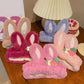 Fluffy bunny headbands in various colors on a wooden surface with a lamp and cake in the background.