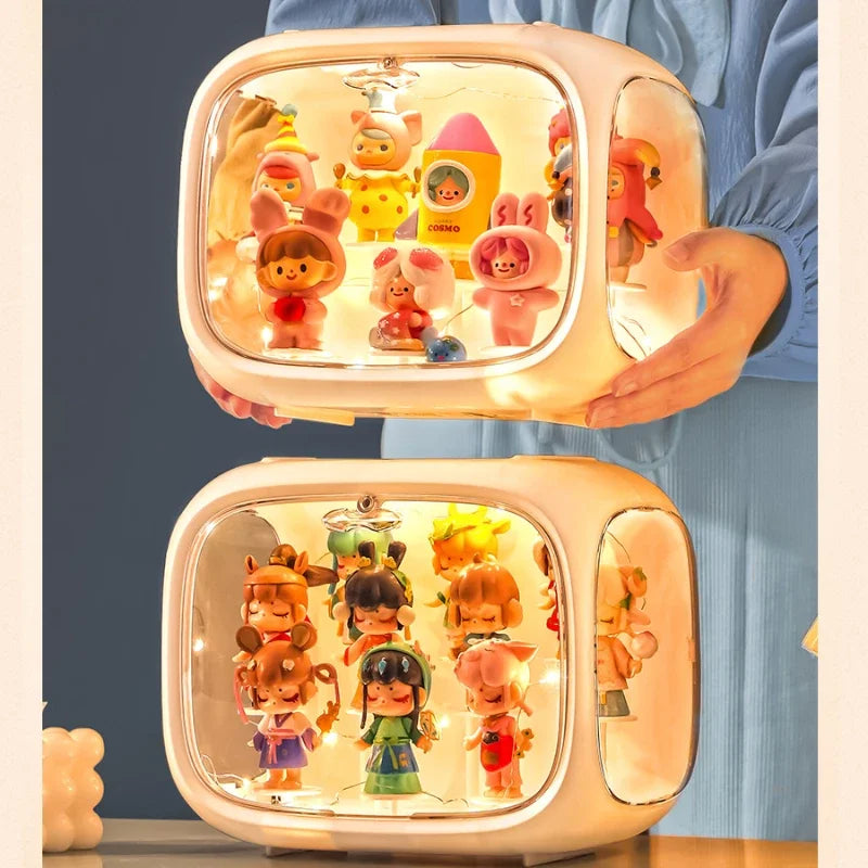 A person in a blue top holds two illuminated cream-colored display boxes containing multiple small cartoon figurines. The boxes have rounded rectangular shapes with clear front panels and warm white internal lighting.