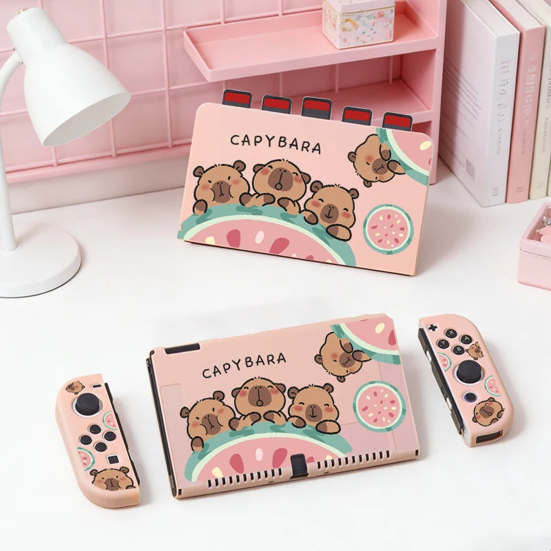 kawaii capybara watermelon switch case, this is the pastel pink version with three capybaras holding onto a watermelon while displaying different happy expressions on their faces,it is also showing a pastel pink dockcase with the same design holding game cards, this is shown on a white desk.
