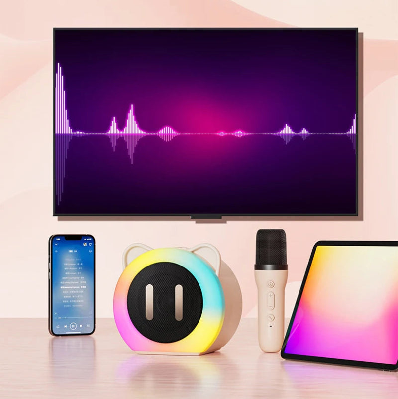 Cute Cat Mini Karaoke Speaker pink edition displayed on a pink table top with a microphone and phone next to it  along with a tv in the background displaying it is compatible with most devices.