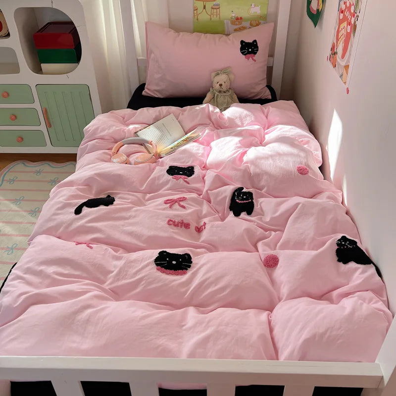 A pink quilted bedding set with black cat embroidery and bows, styled on a white single bed in a child's bedroom.