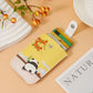 A yellow card holder wallet with a cartoon panda and orange berries design, shown holding several credit cards on a peach-colored background with decorative props.