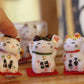 Three small ceramic cat figurines with Japanese text on a wooden surface, branded 'Wontive'.