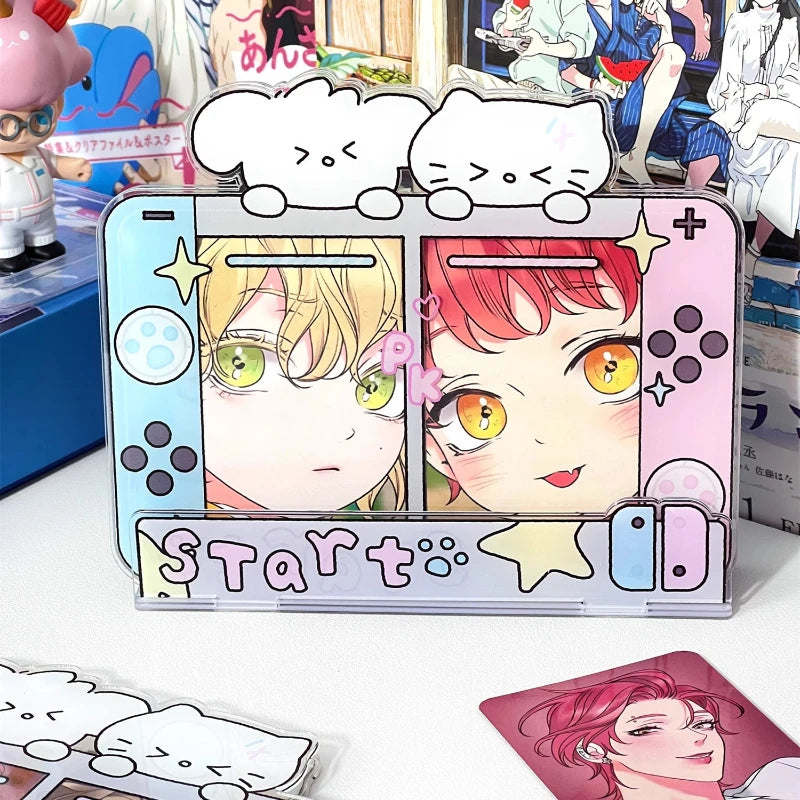 A decorative acrylic desk stand shaped like a retro game console, featuring anime character artwork on the screen and a pink 'START' banner at the bottom. The stand is photographed on a tabletop with other printed cards or stickers nearby and anime figures/posters in the background.