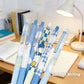 Six blue cartoon gel pens with penguin illustrations are arranged on a wooden desk next to notebooks and a lit desk lamp. The pens feature light blue, navy blue, and white colors with cute penguin designs.