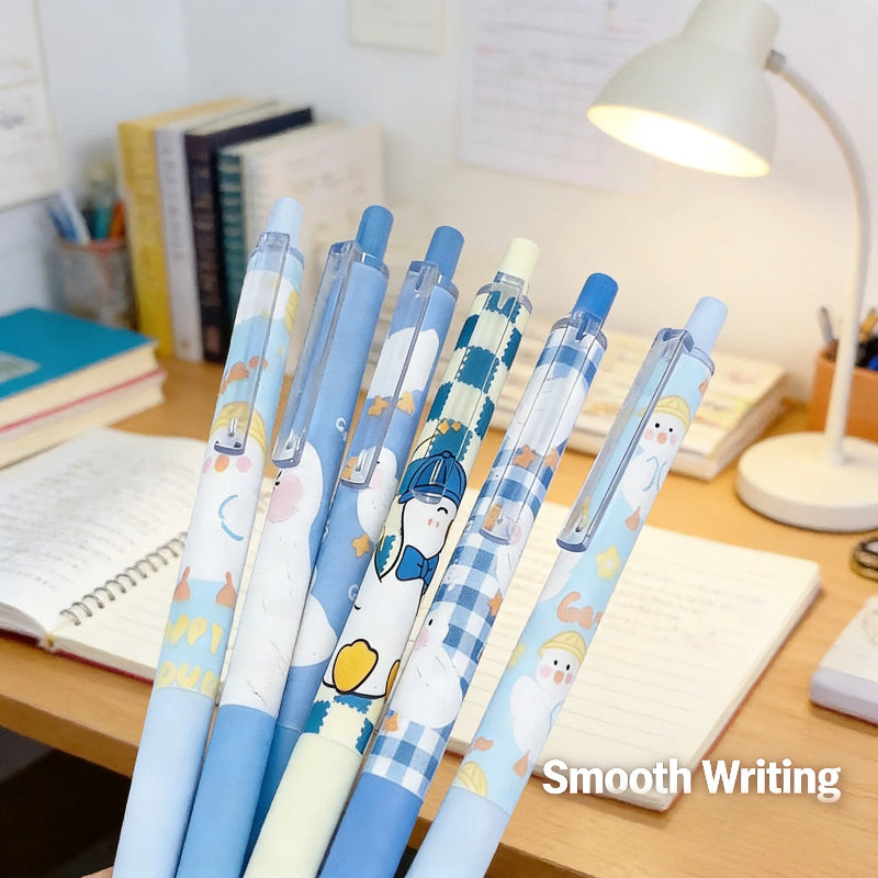 Six blue cartoon gel pens with penguin illustrations are arranged on a wooden desk next to notebooks and a lit desk lamp. The pens feature light blue, navy blue, and white colors with cute penguin designs.