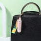 Pink fan with colorful strap attached to a black handbag against a light green background