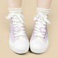 A pair of high-top canvas sneakers in light purple and white, featuring white laces, white rubber soles, and embroidered white flowers with purple centers on the sides. The shoes are shown being worn with white ruffled socks against a solid beige background.