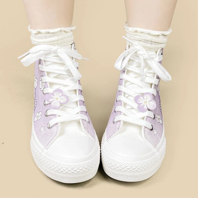 A pair of high-top canvas sneakers in light purple and white, featuring white laces, white rubber soles, and embroidered white flowers with purple centers on the sides. The shoes are shown being worn with white ruffled socks against a solid beige background.