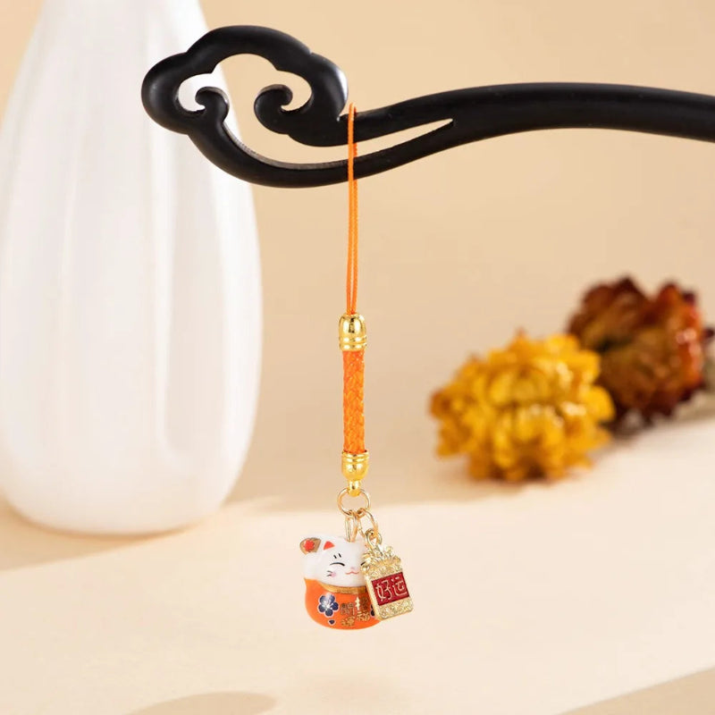 A small hanging charm with an orange cord, gold-toned beads, and a decorative pouch featuring a white cat and Chinese characters, displayed on a black ornamental stand against a warm beige background.