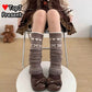 A person seated on a chair wearing long brown knit leg warmers with white heart and bow patterns, paired with dark brown bow shoes. The image includes a 'TopJ Present' logo overlay in the top-left corner and a small teddy bear prop on the floor in the background.