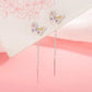 A pair of silver butterfly dangle earrings with pastel purple and yellow rhinestone accents, displayed on a white card over a pink background.