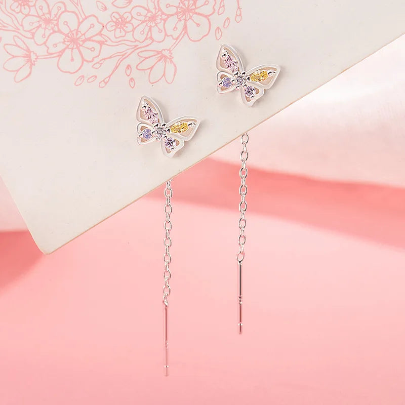 A pair of silver butterfly dangle earrings with pastel purple and yellow rhinestone accents, displayed on a white card over a pink background.